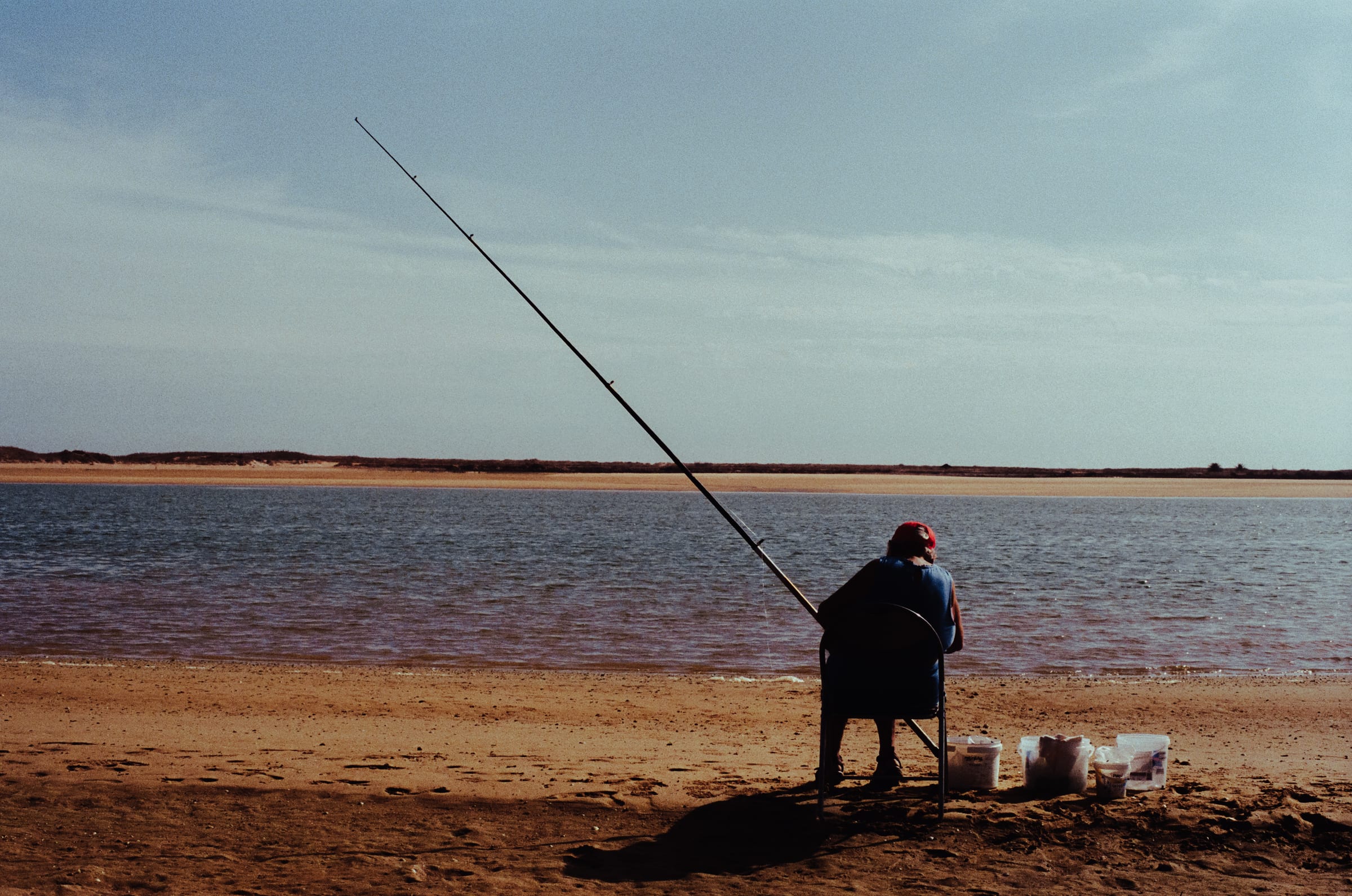 De dos, un pêcheur assis face à la lagune, sa longue canne en diagonale, des seaux à ses pieds.