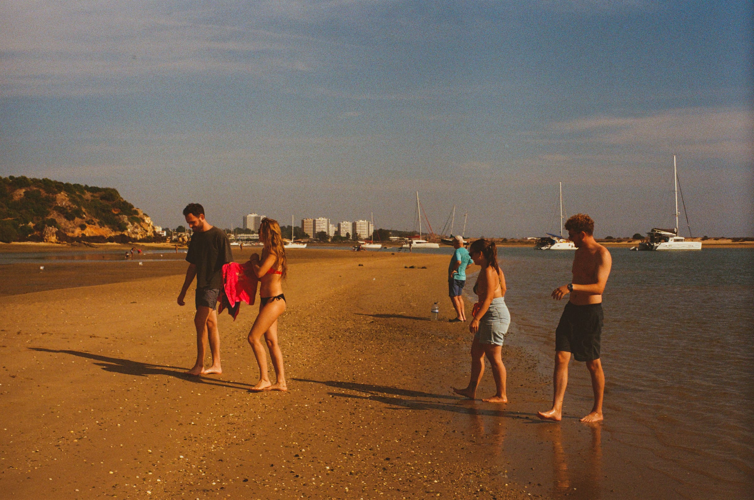 Les copains marchent pieds nus sur le banc de sable, des voiliers et des immeubles au fond.